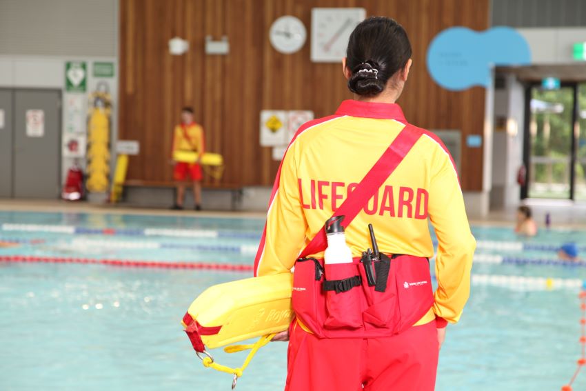 A lifeguard, wearing a bright red and yellow uniform, stands with her back to the camera as she watches swimmers in the pool.