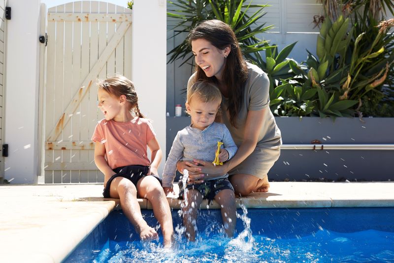 A mother and her two children sit by the side of their backyard swimming pool. The children kick and splash the water and the mother is laughing.