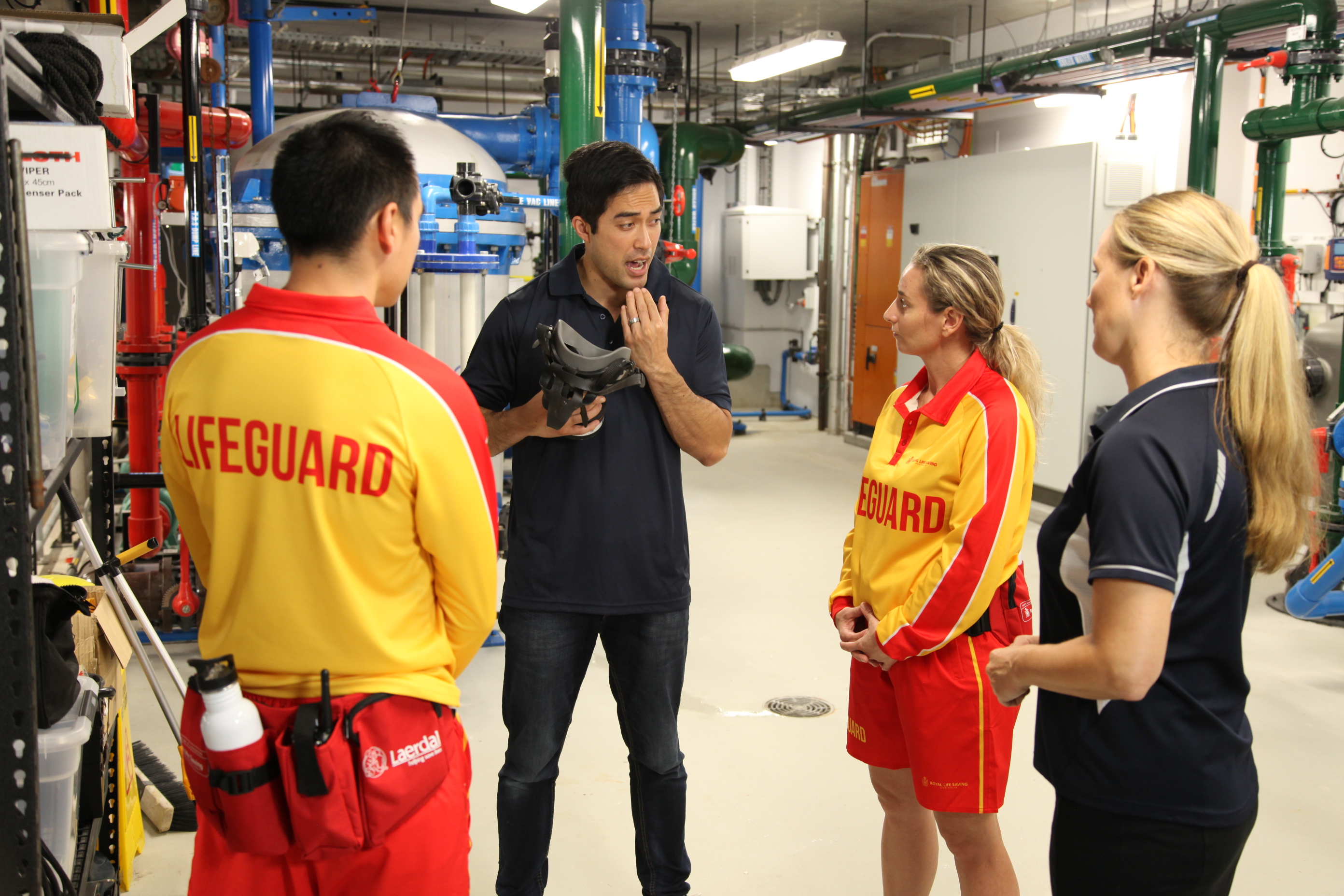 A duty manager is talking to three lifeguards in the pump room of a swimming pool. Behind him are the chlorine tanks and machines for operating the pool.