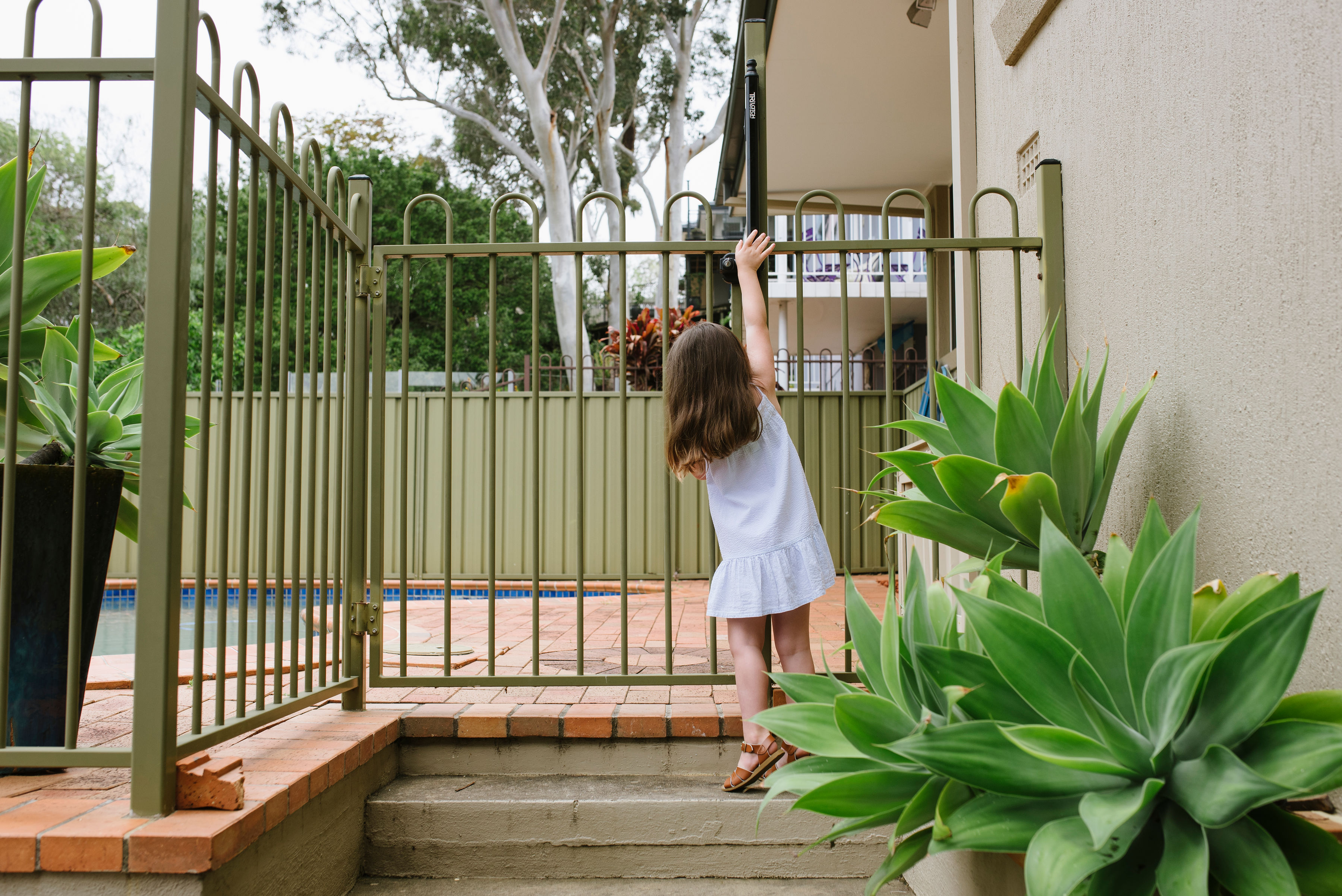 A small child stands in front of a swimming pool fence. She is reaching up to the gate and we can see the blue water of the swimming pool in front of her..