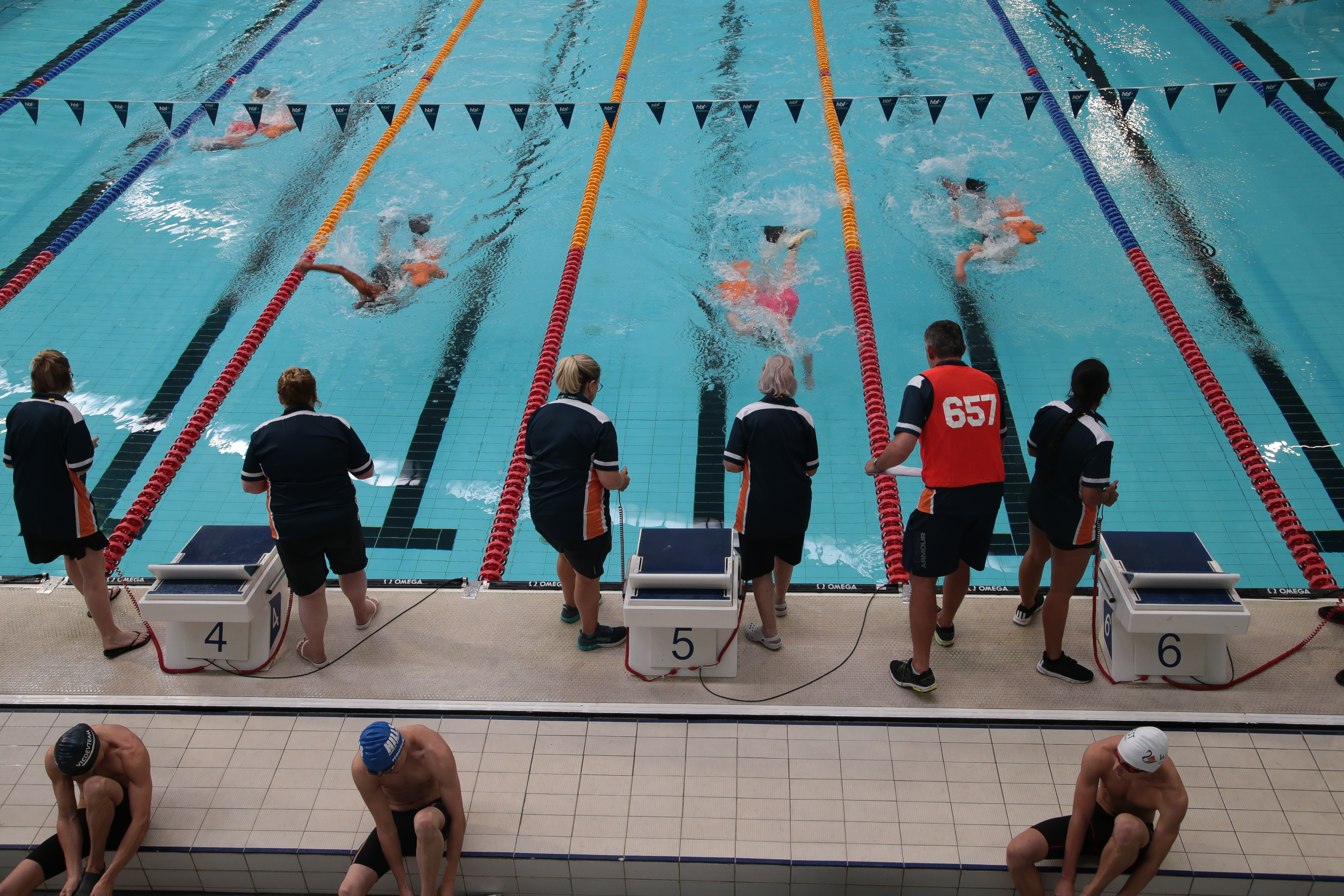 An overhead shot of the end of a swimming pool. Swimmers are closing in on the end of their race, as volunteer time keepers lean over the edge of the pool poised ready to stop their stopwatches.